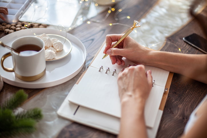 Young woman drinking tea and writing plans or goals for New Year 2021 while her daughter crafting Christmas trees from paper cone, yarns and buttons with placed stars and fairy lights on wooden table.